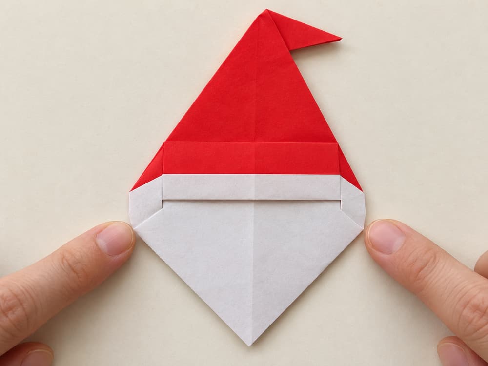 origami santa step 8: turn the face forward and fold the very top of the red hat sideways, hands visible