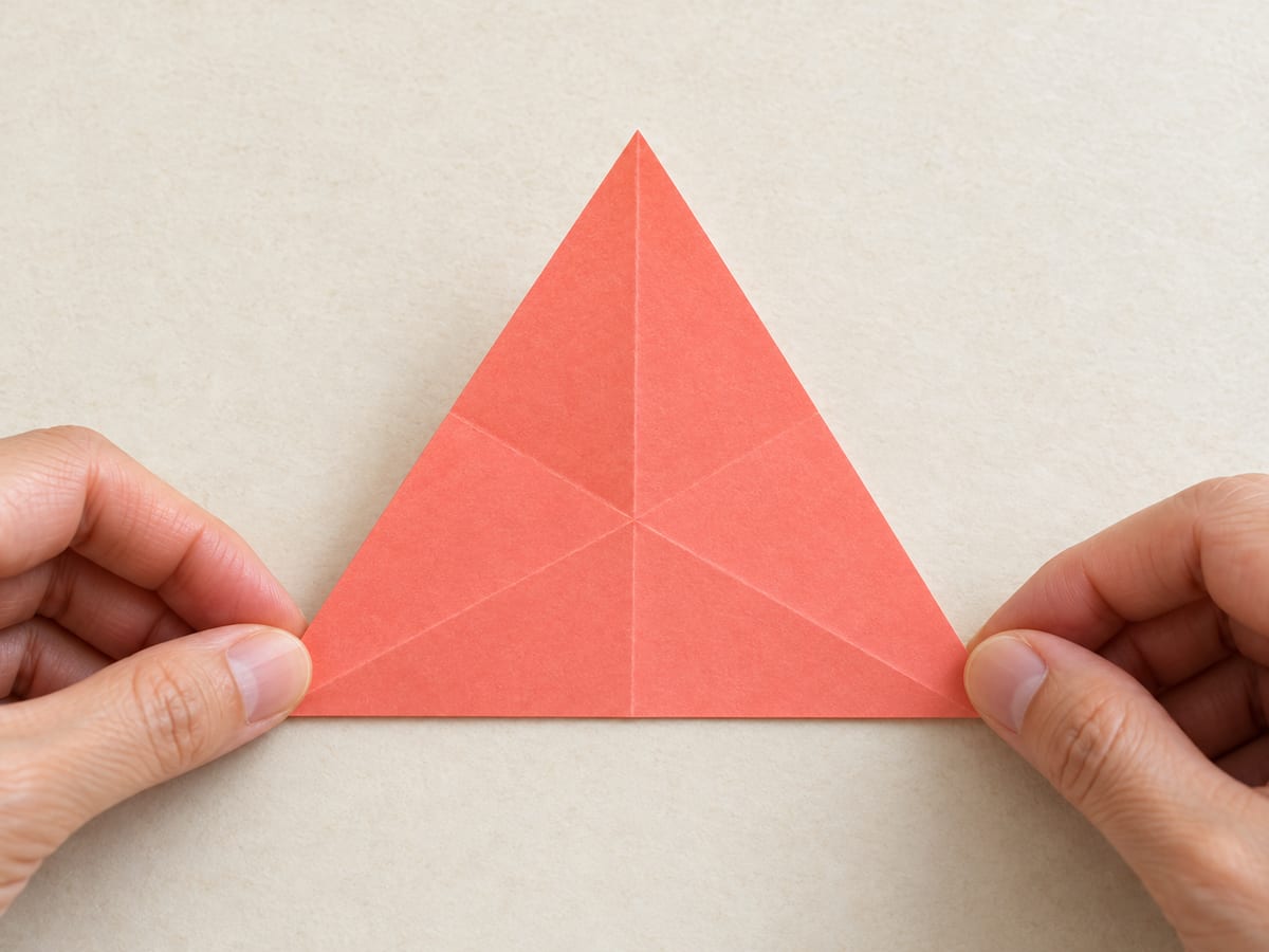 origami star step 6: fold the triangle in half corner to corner, then reopen; repeat using each corner to mark three center guides, hands visible
