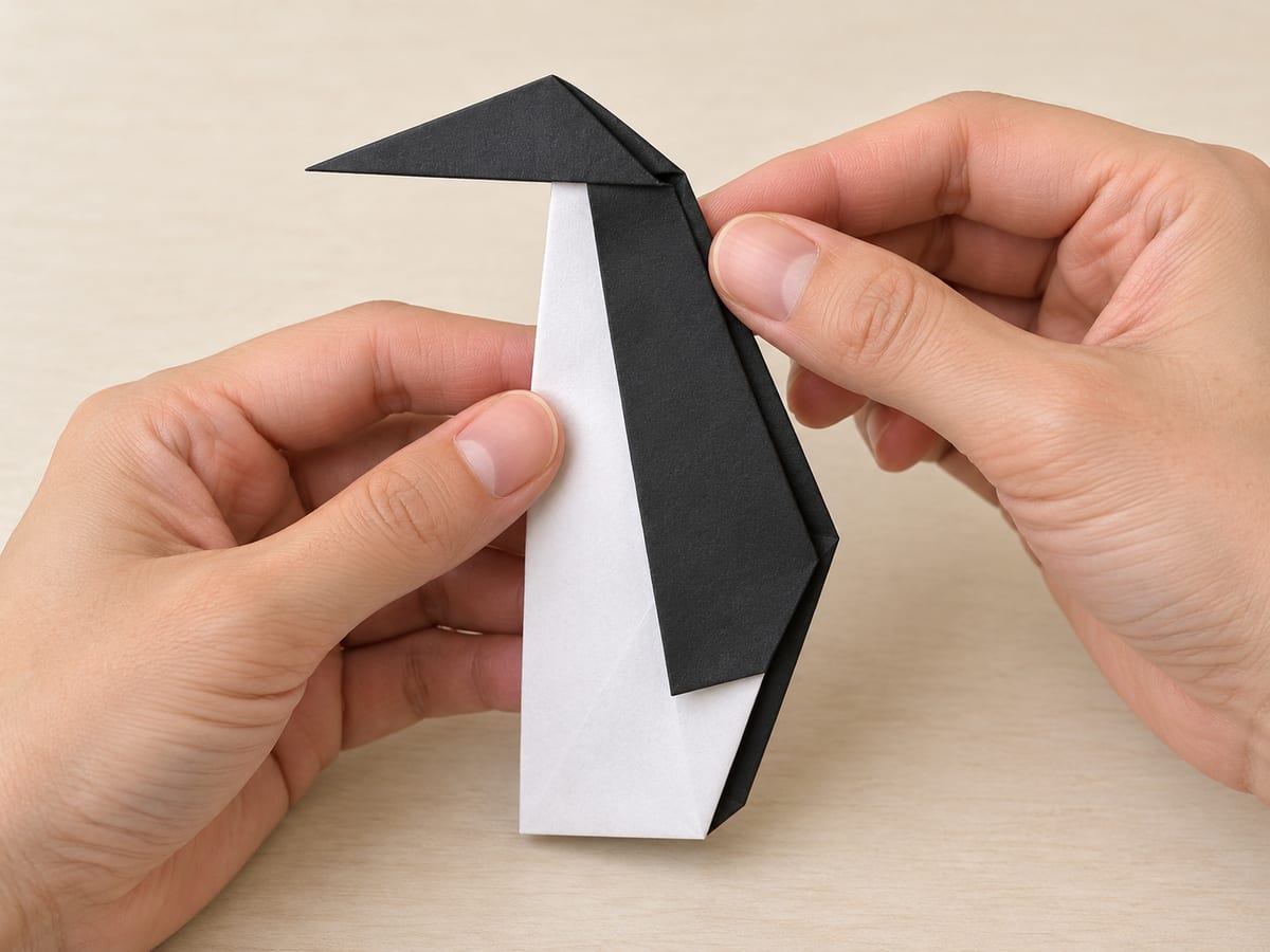 origami penguin step 8: open the neck layers slightly, reverse the head outward along its crease, and close the body around it, hands visible