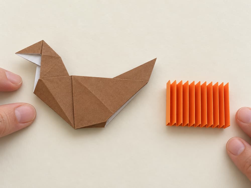 origami turkey step 9: accordion-fold the orange square into equal half-inch strips, alternating forward and backward folds across the whole sheet, hands visible