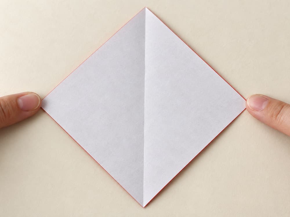origami santa step 1: start white side up with a point facing down; fold left point to right point and reopen, hands visible