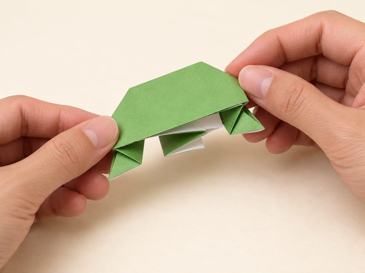 origami frog step 11: turn the model over and place the folded spring underneath its body, hands visible