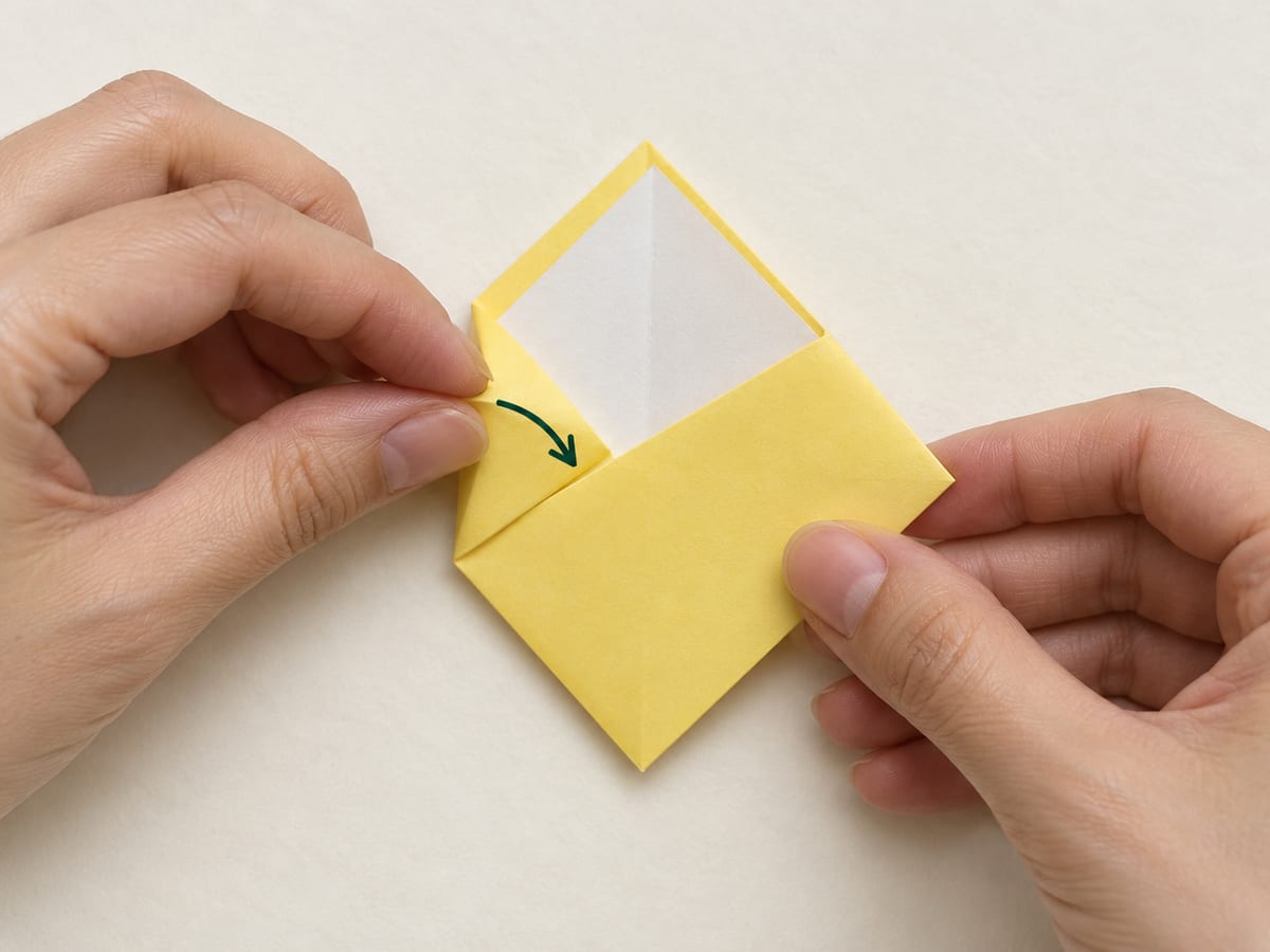 origami bookmark step 6: lift and tuck the left corner into the same pocket, keeping the outer edges aligned, hands visible