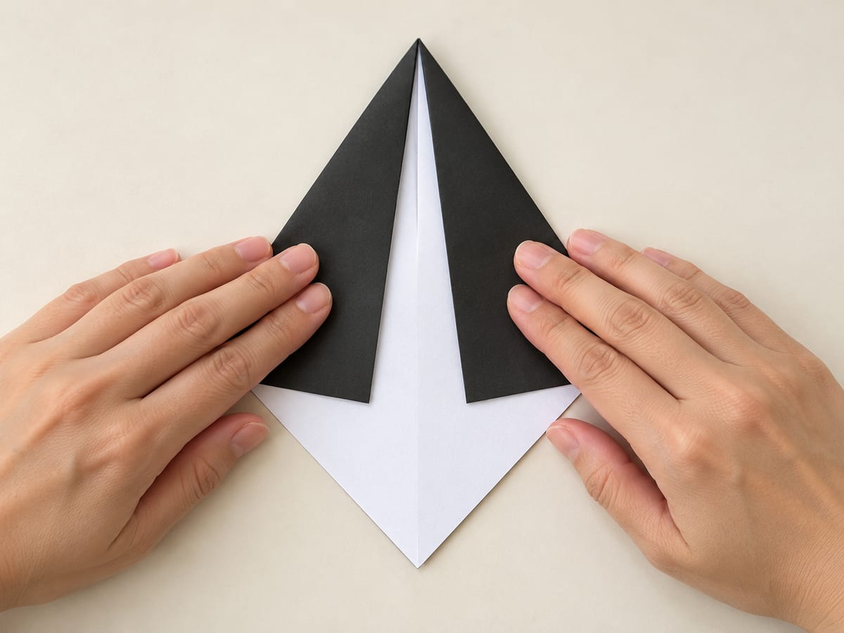 origami penguin step 2: fold both upper sloping edges inward, leaving a narrow gap between them instead of meeting at the center, hands visible