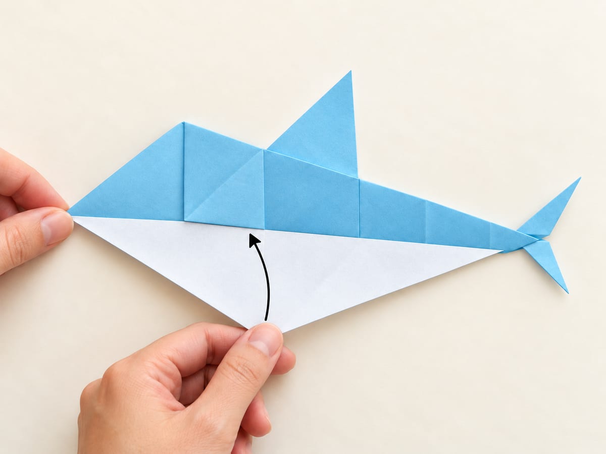 origami shark step 11: fold the lower half behind the upper half and keep the dorsal fin above the body, hands visible