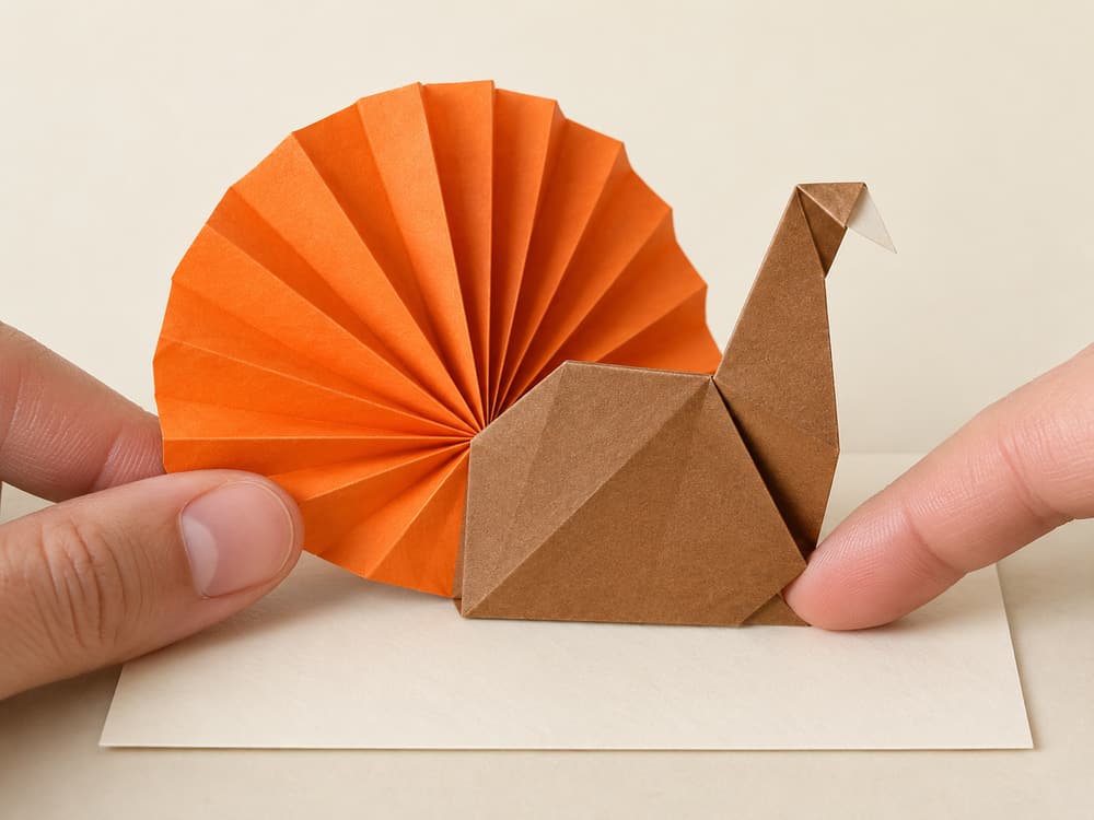 origami turkey step 12: glue the fan behind the body; hold until set, then lean the turkey against a place card, hands visible