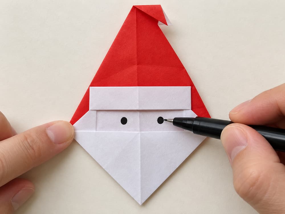 origami santa step 9: draw two small eyes just above the pointed beard and press the side folds flat, hands visible