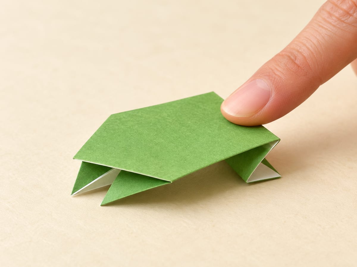 origami frog step 12: press the back gently against a firm table, then slide your finger off to release the spring, hands visible