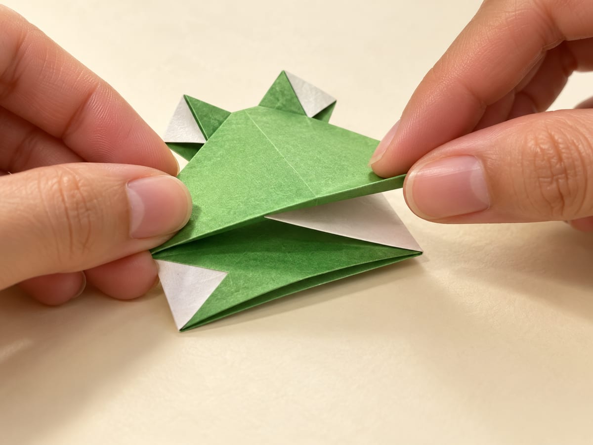 origami frog step 10: fold that same section halfway back toward the bottom edge, forming a Z-shaped spring, hands visible