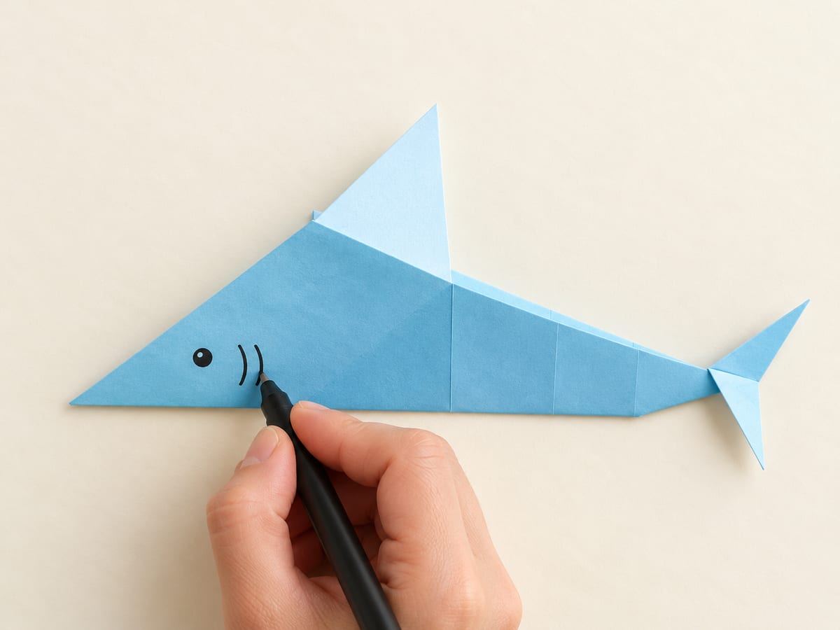 origami shark step 13: draw a small eye and curved gill near the broad left head and press the body flat, hands visible