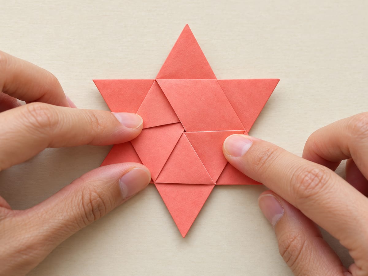 origami star step 12: slide the last corner beneath the first pleat and press the center flat to finish the six-point star, hands visible