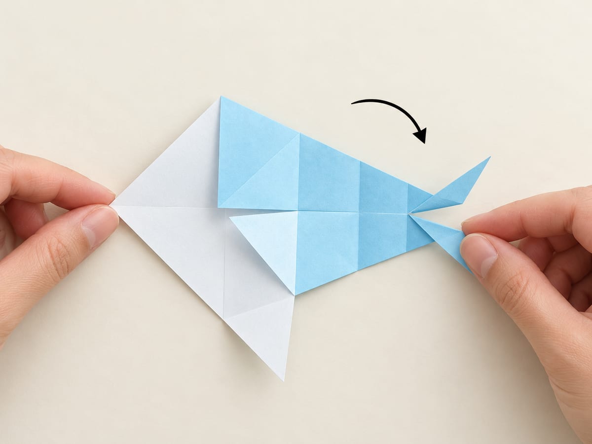 origami shark step 10: rotate the paper clockwise one quarter turn with the split tail pointing right, hands visible