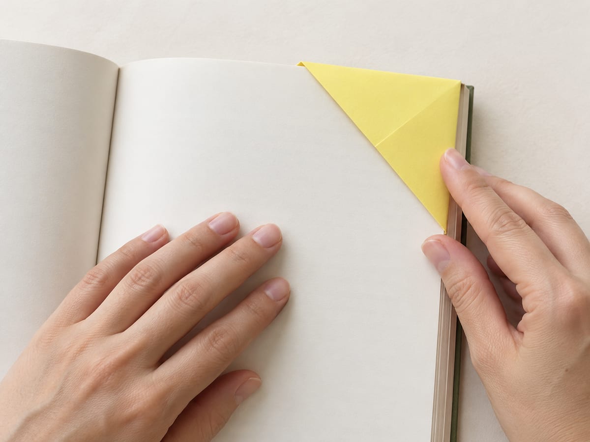 origami bookmark step 7: flatten the pocket and slip it over a page corner, keeping the paper clear of the text, hands visible